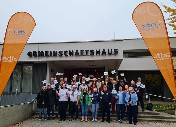 Eine große Gruppe Menschen steht vor einem Gebäude. Die Menschen tragen überwiegend Winterkleidung. Einige von Ihnen schwenken Fahnen, auf denen das Logo des Jungen ADFC zu sehen ist. Auf dem Gebäude steht "Gemeinschaftshaus". Am linken und rechten Bildrand sind orange Flaggen aufgestellt, die das Logo des ADFC zeigen.